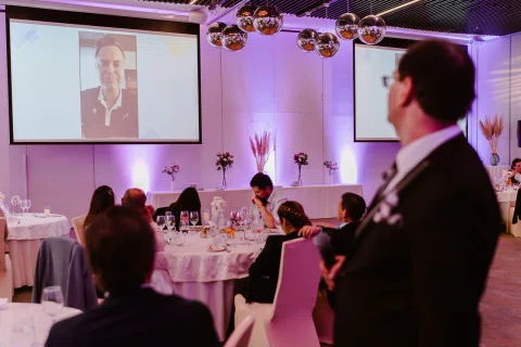 Varias personas observan una proyección en una pantalla dentro de un salón, con un hombre de traje oscuro en primer plano.