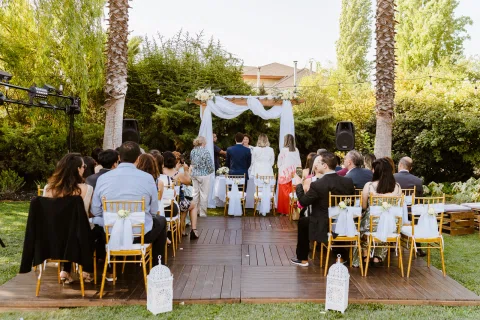 Grupo de personas sentadas frente a una estructura de madera decorada con telas blancas y flores en un jardín.
