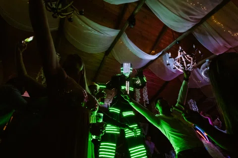 Varias personas con trajes iluminados en verde levantan los brazos entre asistentes de la fiesta.