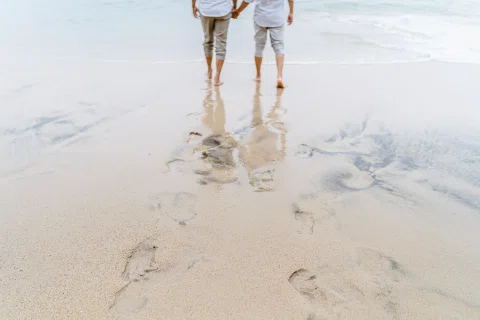 Piernas de dos personas caminando de espaldas y tomadas de la mano, reflejadas sobre la arena mojada junto al mar.