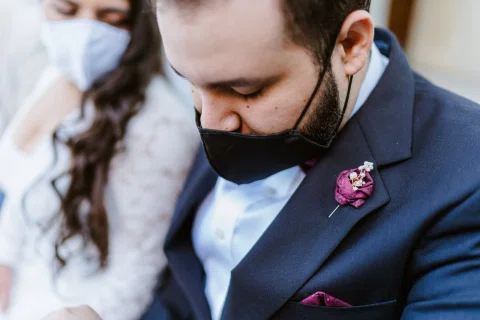 Primer plano de un hombre con traje oscuro y mascarilla, con una flor burdeos en la solapa.