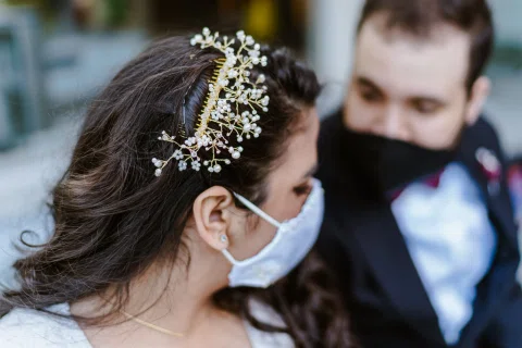 Primer plano de una mujer con mascarilla y pequeñas flores en el cabello, con un hombre desenfocado detrás.