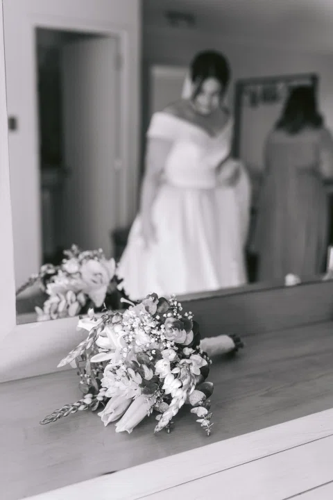 Ramo de flores descansa en primer plano frente al reflejo de una mujer con vestido blanco, en blanco y negro.
