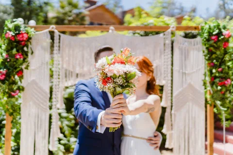 Hombre con traje azul y mujer con vestido blanco sostienen un ramo de flores hacia la cámara frente a una estructura decorada.