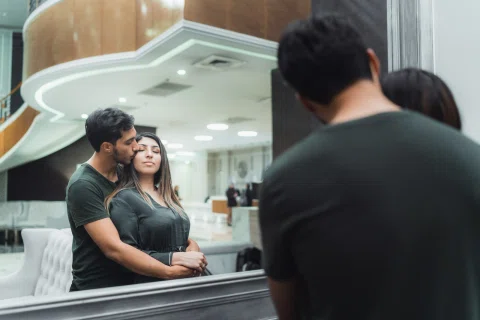El reflejo en un espejo muestra a un hombre abrazando y besando la mejilla de una mujer vestida de oscuro.