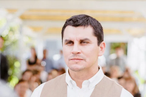 Retrato de un hombre con chaleco beige y camisa blanca, con varias personas desenfocadas al fondo.