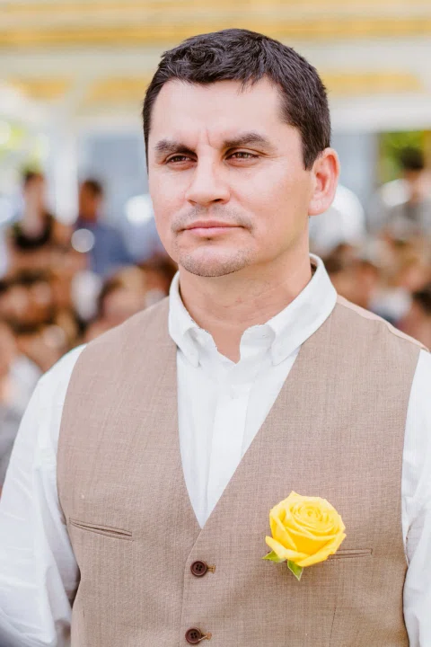 Retrato de un hombre con chaleco beige y una flor amarilla prendida al pecho.