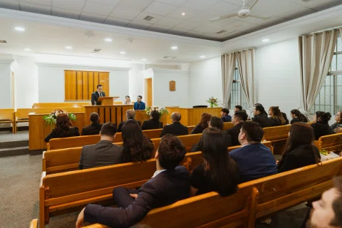 Vista amplia de una sala con personas sentadas en bancos y un atril de madera al fondo.