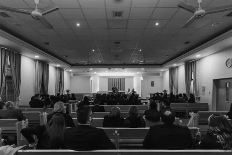 Vista amplia en blanco y negro de una sala con bancos ocupados y un atril al fondo.