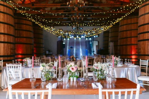 Salón con grandes barriles de madera, mesas decoradas y guirnaldas de luces.