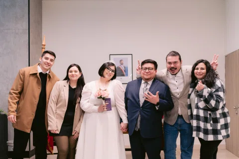 Seis personas posan juntas en un espacio interior; una mujer viste de blanco y un hombre lleva traje oscuro.
