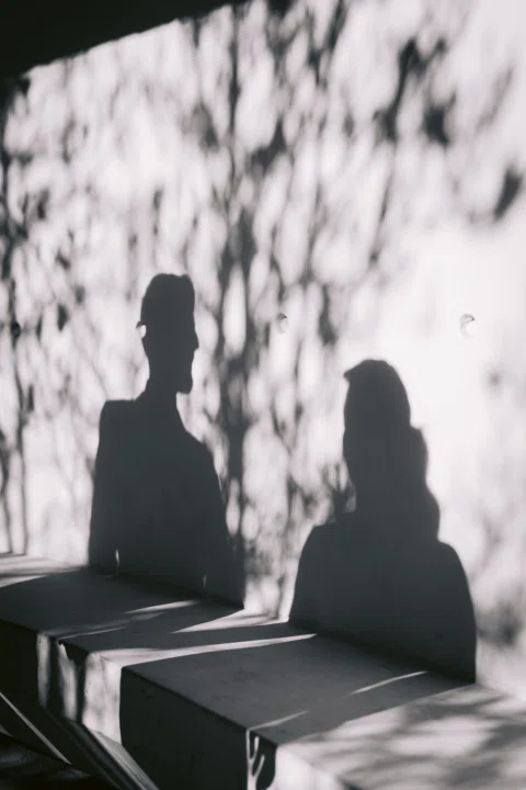 Sombras de un hombre y una mujer proyectadas sobre un muro y una banca, en una fotografía en blanco y negro.