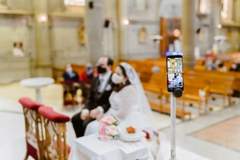 Teléfono móvil montado en un soporte frente a una mujer con vestido blanco y un hombre con traje oscuro sentados dentro de una iglesia.