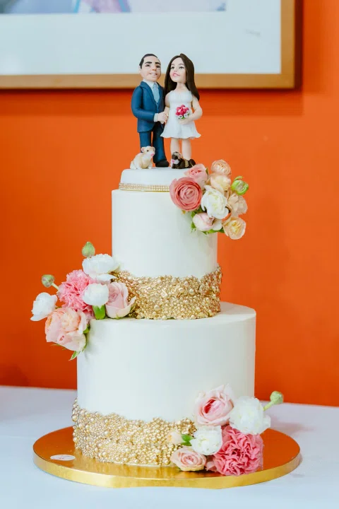 Torta de tres niveles decorada con flores rosadas frente a una pared naranja.