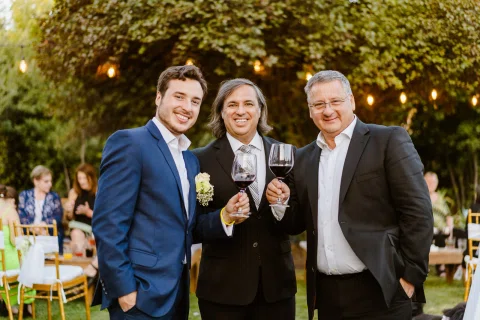 Tres hombres posan juntos al aire libre sosteniendo copas de vino tinto.