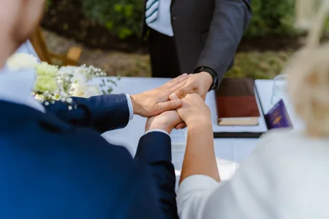 Detalle de tres manos unidas sobre una mesa, con un ramo blanco desenfocado al fondo.