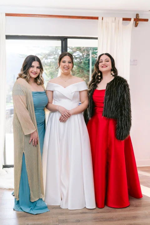 Tres mujeres posan juntas; la del centro lleva vestido blanco y las otras visten azul y rojo.