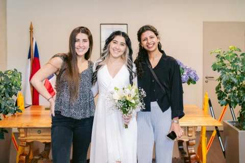 Tres mujeres posan juntas; la mujer del centro lleva vestido blanco y sostiene un ramo.