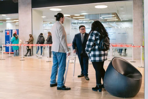 Tres personas conversan en un área interior de atención con divisiones de vidrio y asientos.