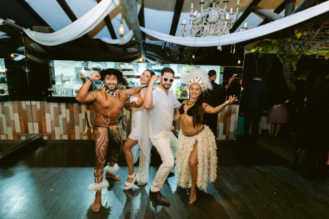 Tres personas posan y bailan frente a una barra, dos con accesorios de fiesta.
