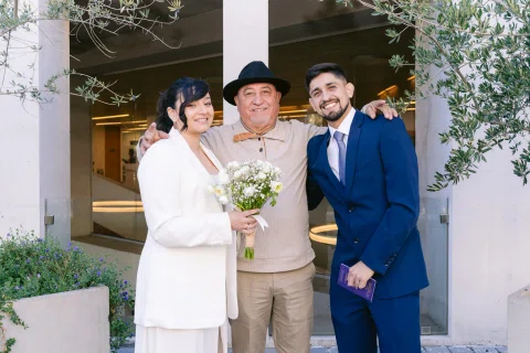 Mujer con traje blanco y ramo y hombre con traje azul posan junto a un hombre con sombrero al aire libre.