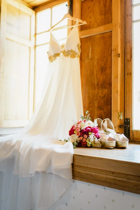 Vestido blanco colgado junto a una ventana con un ramo de flores apoyado cerca.