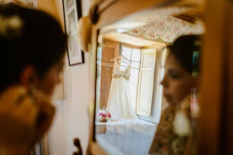 Vestido blanco colgado junto a una ventana y ramo de flores reflejados en un espejo, con dos personas desenfocadas a los lados.