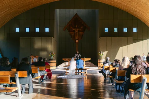 Vista amplia del interior de una capilla con personas sentadas y dos personas al frente.