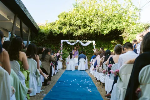 Vista general de un pasillo con alfombra azul, personas sentadas a ambos lados y un arco floral al fondo.